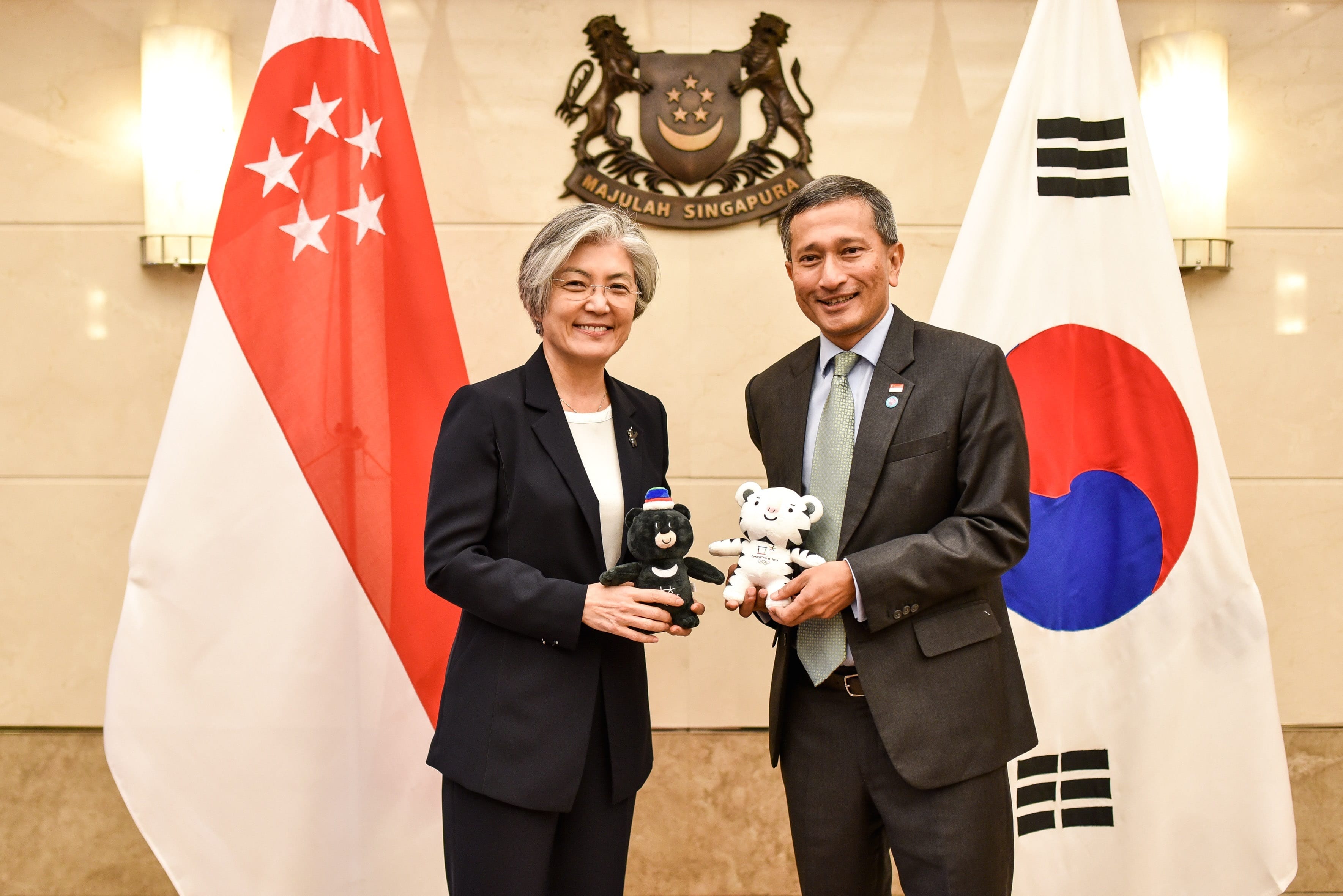 Two people holding PyeongChang Olympic mascots with Singapore and South Korean flags in background.